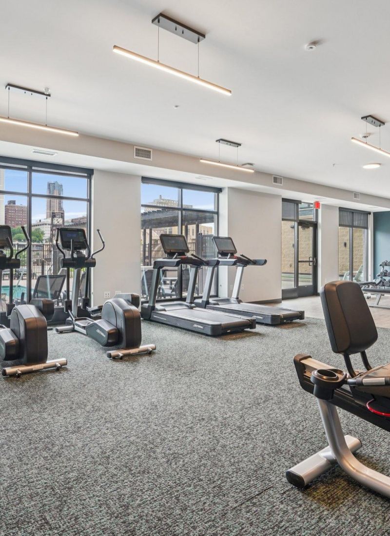 The Scenic modern gym with large windows, treadmills, stationary bikes, and other exercise equipment on a carpeted floor.