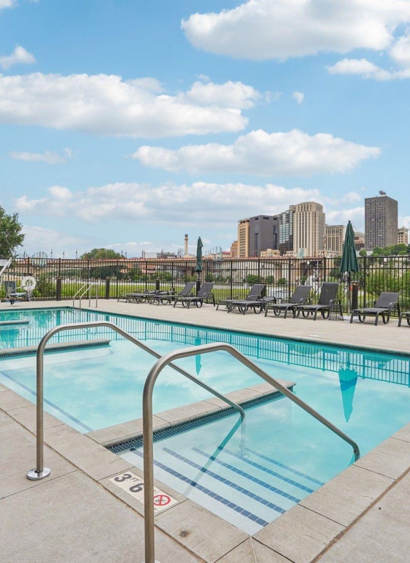 The Scenic outdoor swimming pool with metal handrails, surrounded by city buildings and blue sky with clouds.