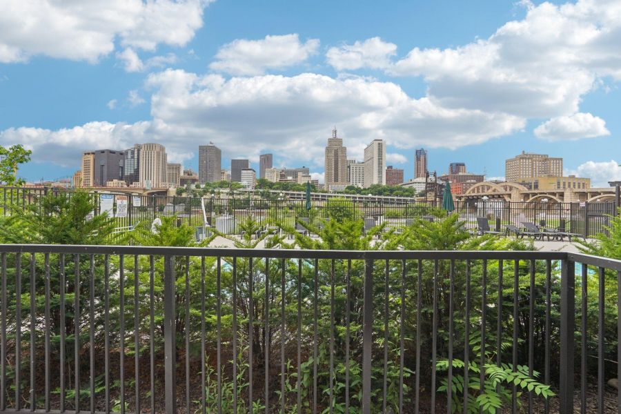 The Scenic city skyline with tall buildings behind a metal fence and green plants under a partly cloudy sky.