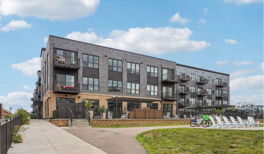 The Scenic modern three-story apartment building with balconies, bike racks, and a sidewalk under a blue sky with clouds.