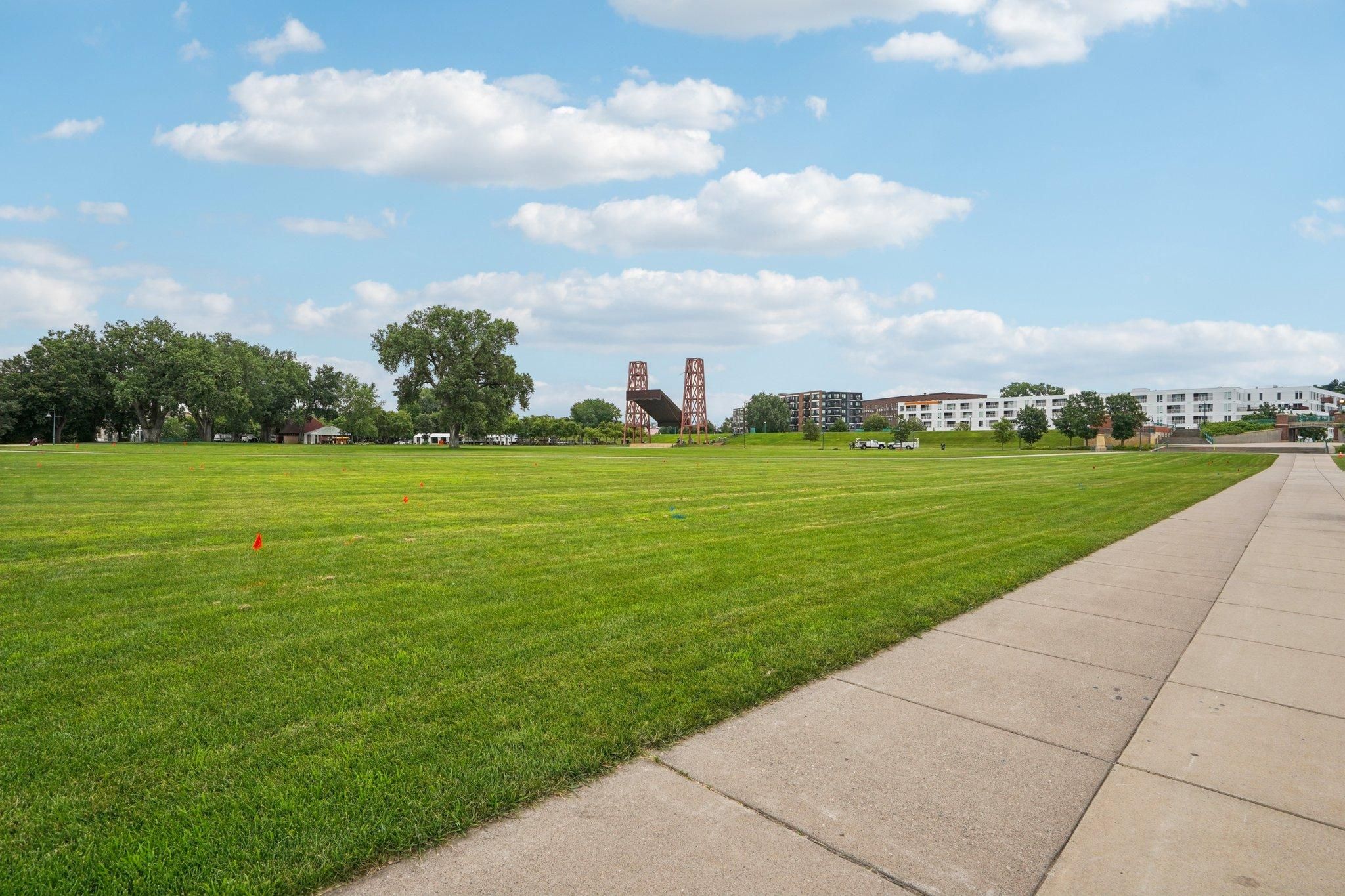 The Scenic a wide grassy field with a sidewalk, trees, and buildings under a partly cloudy sky.